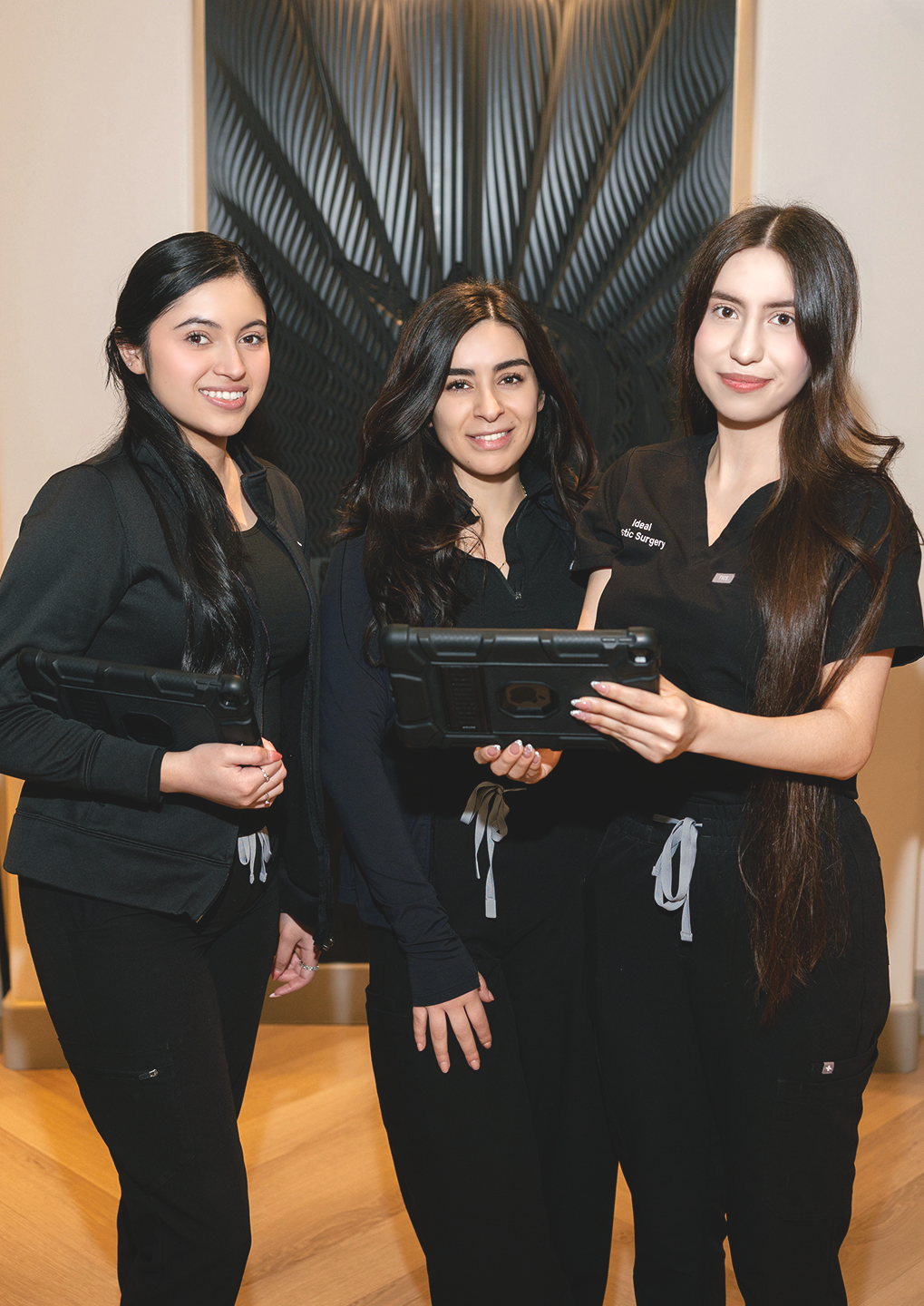 Three female staff members in black uniforms stand together in a clinic lobby, each holding a tablet.