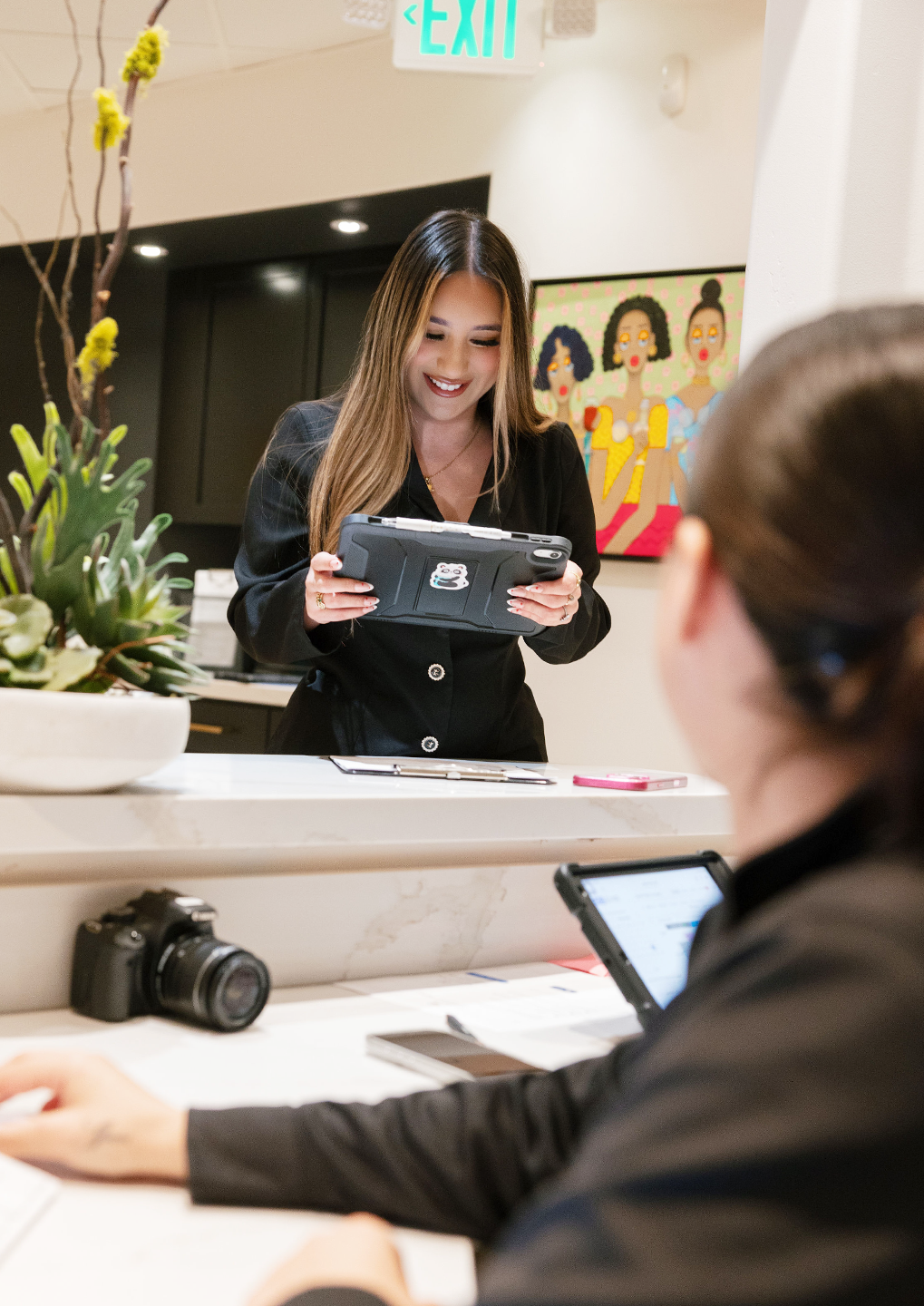 Receptionist behind a white marble counter smiles while showing a tablet to a customer across the desk, indoors with plants nearby.